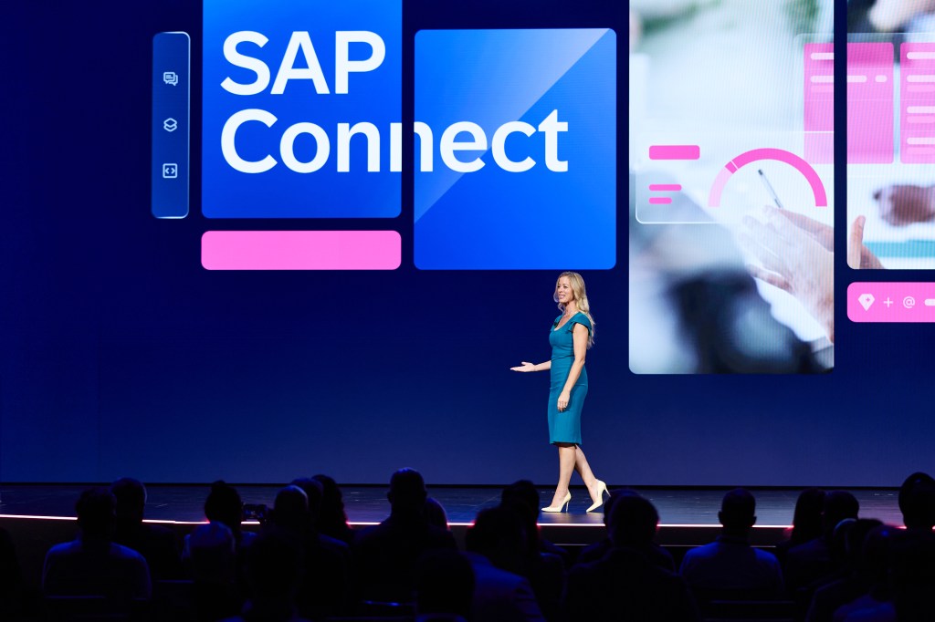 Presentadora en un escenario con una pantalla que dice 'SAP Connect', rodeada de gráficos y con un público visible.