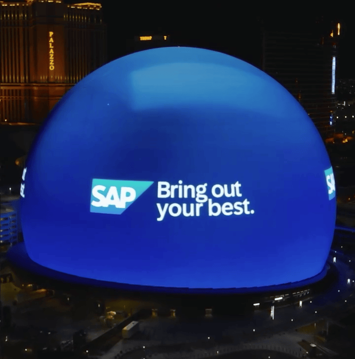 Edificio iluminado de noche con un gran domo azul que muestra el logotipo de SAP y el eslogan 'Bring out your best'.